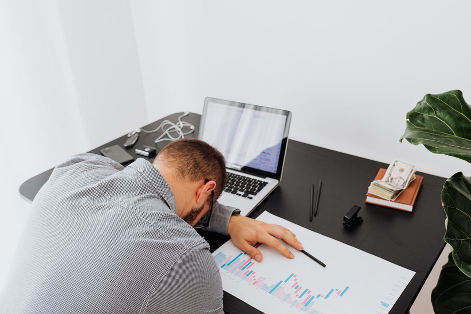 Overworked businessman falling asleep on office desk with laptop and charts, depicting stress and exhaustion.